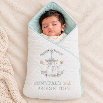 A sleeping newborn baby wrapped in a white quilted swaddle blanket with a light blue hood and pastel floral embroidery, lying on a beige textured blanket.