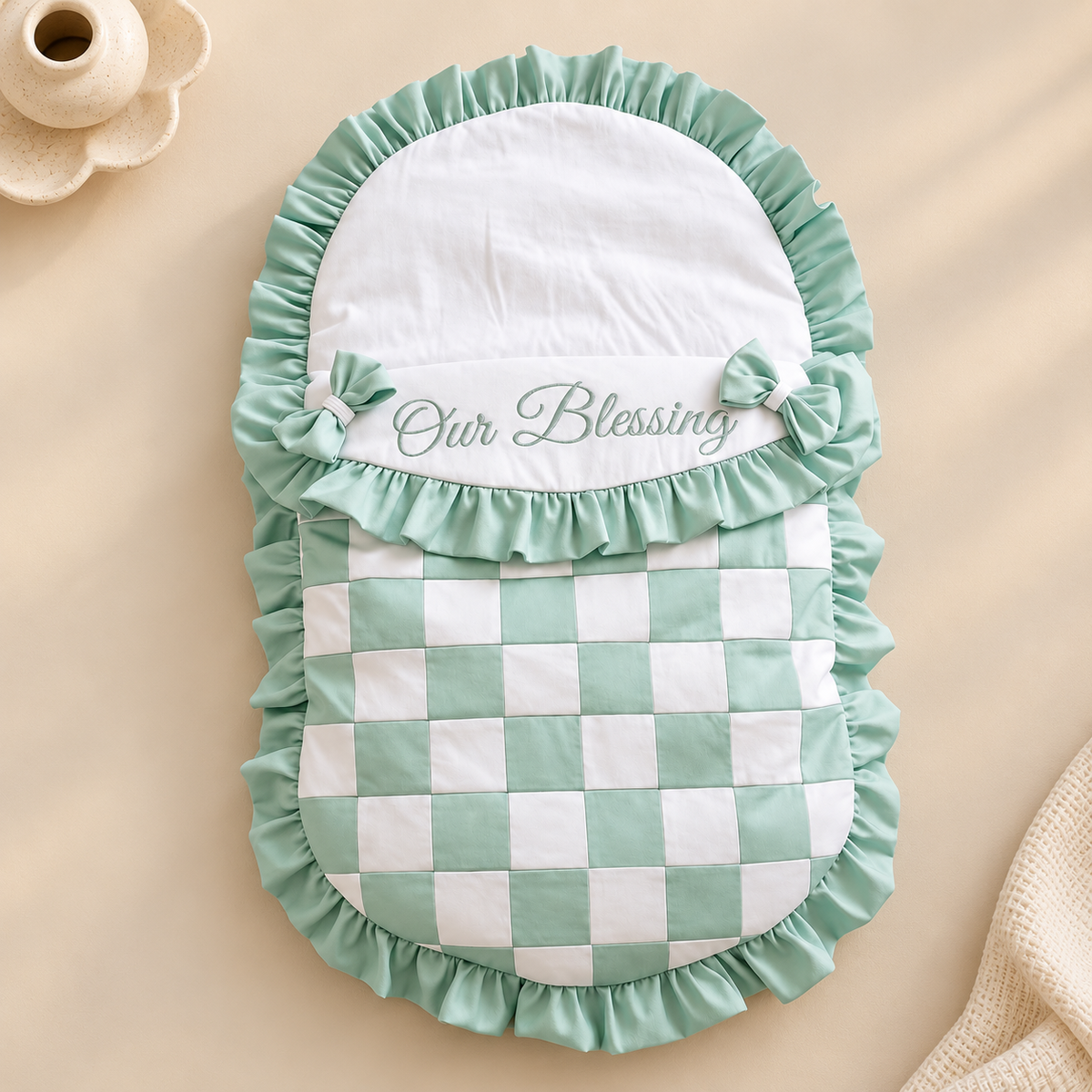 A baby blanket or footmuff with a mint green and white checkered pattern, featuring a white top panel with 'Our Blessing' embroidered in green, ruffled trim, and matching bow details, laid flat on a beige background.