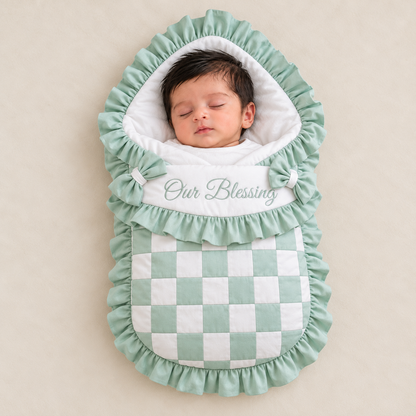 A sleeping newborn baby wrapped in a mint green and white checkered bassinet cover with ruffled edges and the words 'Our Blessing' embroidered on the front, set against a warm beige background.