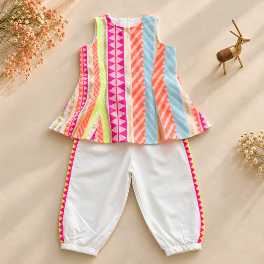 A children's two-piece outfit laid flat on a beige background with decorative dried flowers and a small deer figurine. The set includes a sleeveless top with vertical multicolor striped and geometric panels in pink, orange, yellow, blue, and white, paired with white pants featuring bright pink and yellow striped side trim.