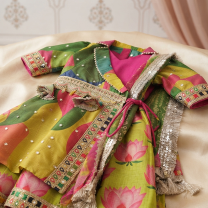 A colorful traditional embroidered garment laid flat on a cream satin surface, featuring bright pink, yellow, green, and orange floral and leaf patterns with metallic silver sequin borders and tassels.
