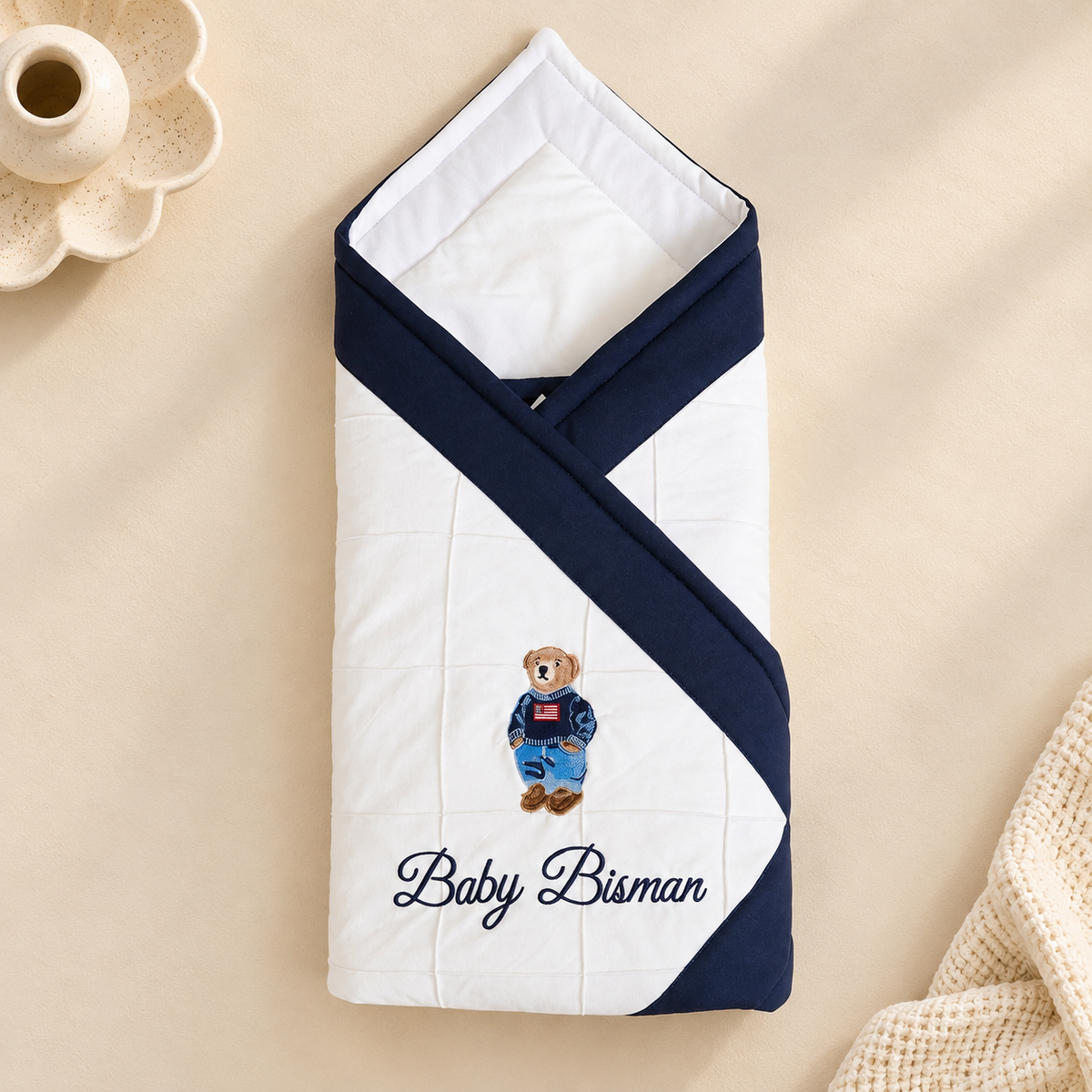 A personalized baby swaddle blanket with a white body, navy blue borders, and an embroidered teddy bear wearing blue overalls. The name 'Baby Bisan' is embroidered in navy blue script at the bottom.