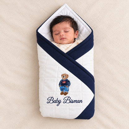 A sleeping newborn baby is swaddled in a custom quilted wrap blanket. The blanket is white with navy blue borders, featuring an embroidered teddy bear and the personalized name 'Baby Bisan' in navy blue script.