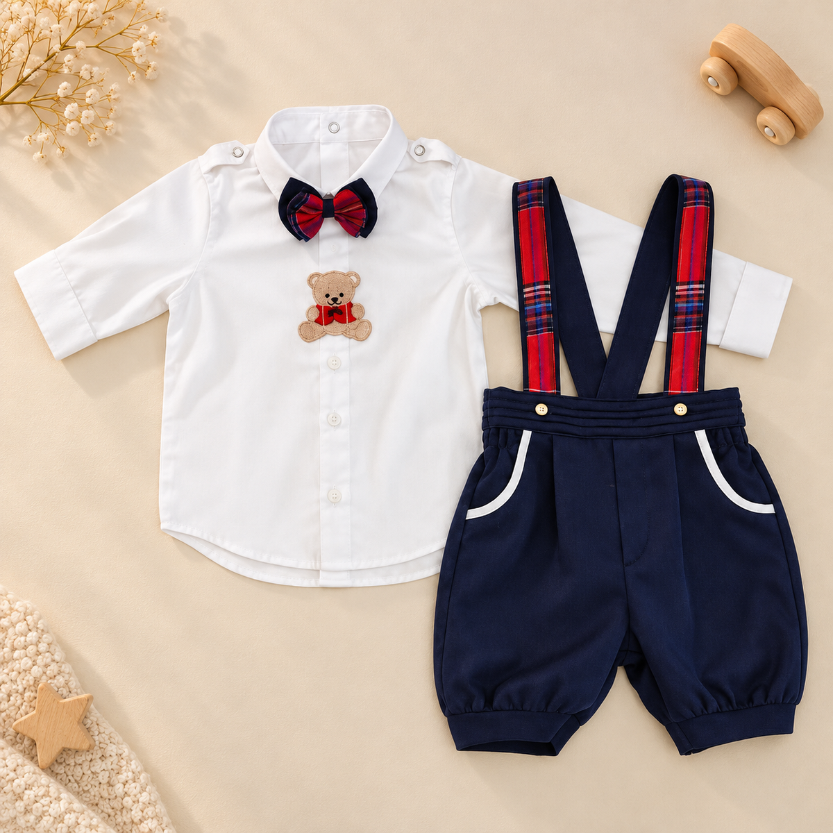 A baby boy's two-piece outfit set featuring a white long-sleeve button-up shirt with a red and navy bow tie and teddy bear patch, paired with navy blue shorts that have red and blue plaid suspenders and white pocket trim, laid flat on a beige background with styled props.