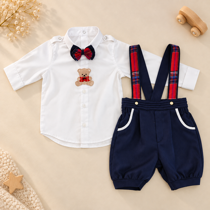 A baby boy's two-piece outfit set featuring a white long-sleeve button-up shirt with a red and navy bow tie and teddy bear patch, paired with navy blue shorts that have red and blue plaid suspenders and white pocket trim, laid flat on a beige background with styled props.