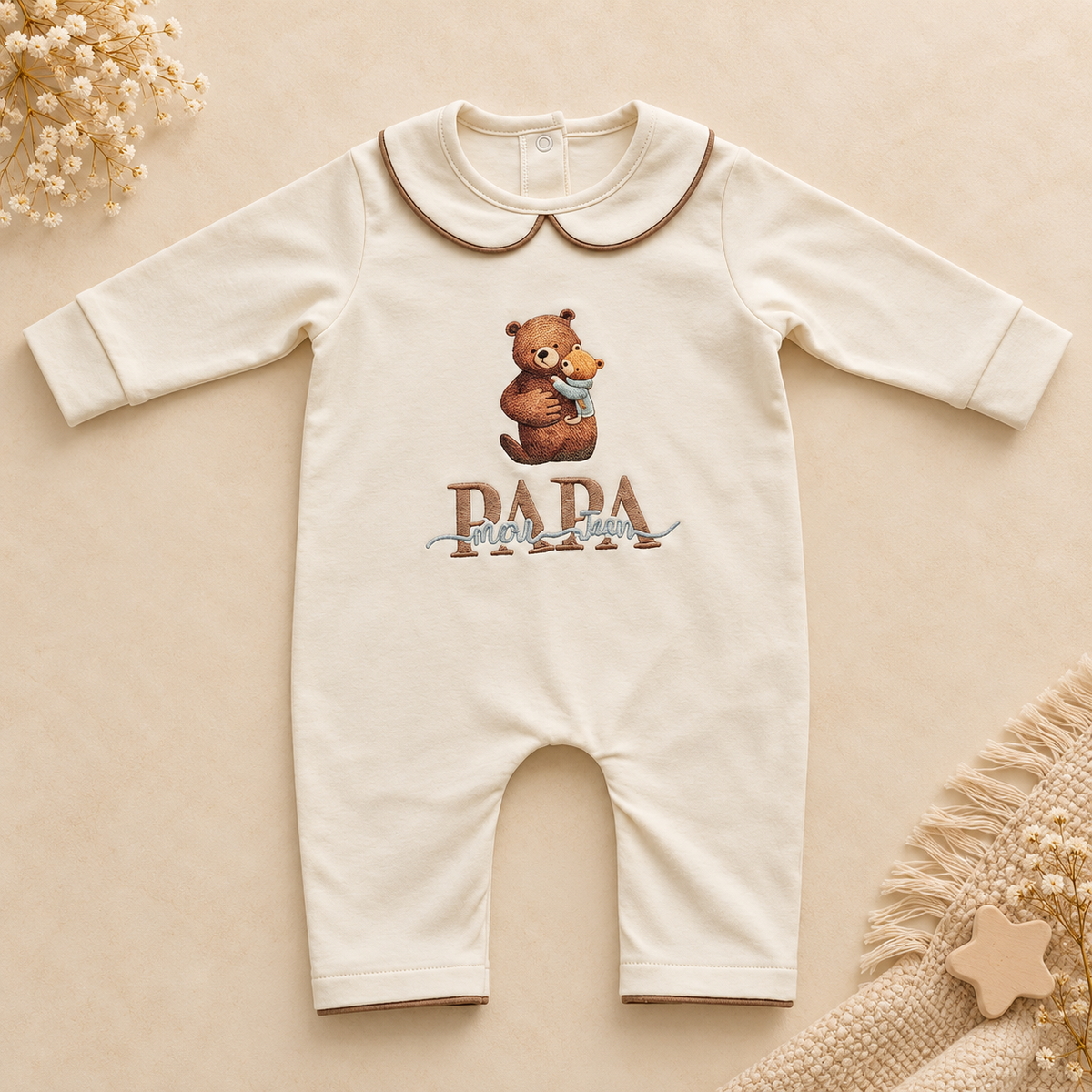 A cream-colored baby long-sleeve romper with a Peter Pan collar, featuring a graphic of a teddy bear holding a small blue bird and the text 'PAPA' below it. The romper is displayed flat on a warm beige background with decorative dried flowers and a knitted textile in the corners.