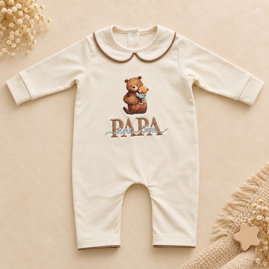A cream-colored baby long-sleeve romper with a Peter Pan collar, featuring a graphic of a teddy bear holding a small blue bird and the text 'PAPA' below it. The romper is displayed flat on a warm beige background with decorative dried flowers and a knitted textile in the corners.