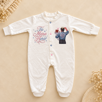A white long-sleeve baby footed sleeper with a button-up front, featuring 'The Mera Hero' text and a graphic of a baby dressed as a hero on the front, laid flat on a beige background with decorative dried flowers.