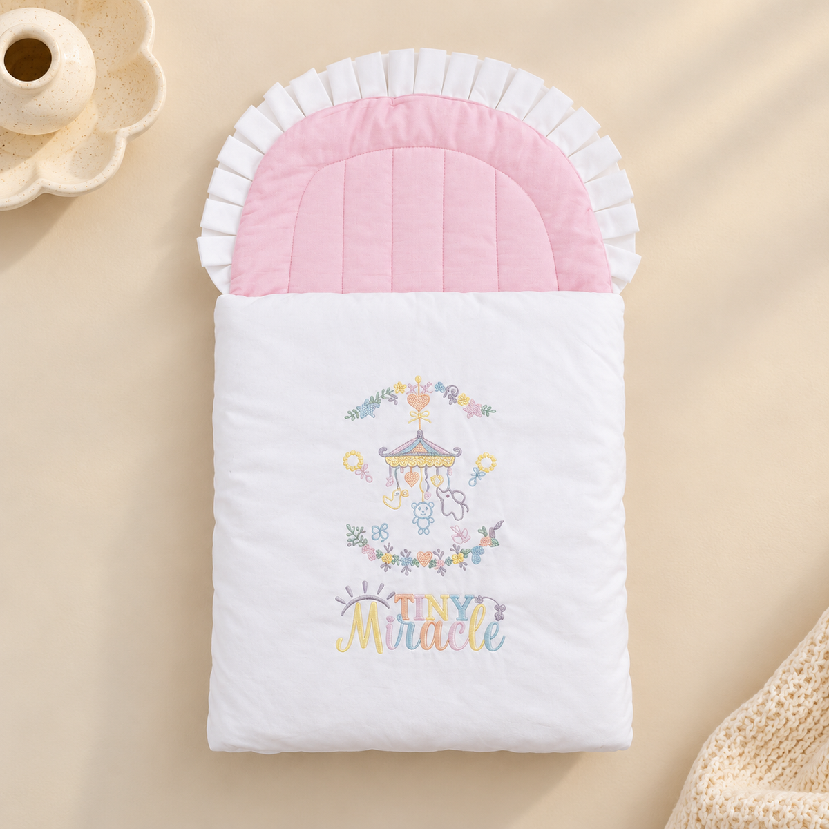 A baby sleep sack or quilted blanket with a white body, pink ruffled trim, and a colorful embroidered carousel and 'Tiny Miracle' text, styled on a beige surface with soft decorative props.