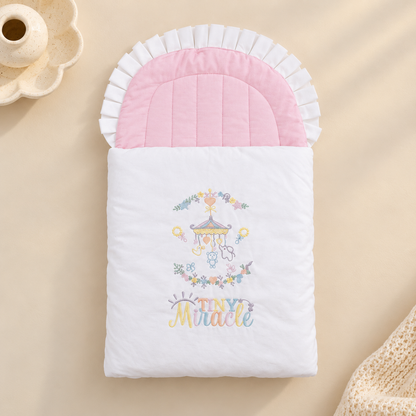 A baby sleep sack or quilted blanket with a white body, pink ruffled trim, and a colorful embroidered carousel and 'Tiny Miracle' text, styled on a beige surface with soft decorative props.