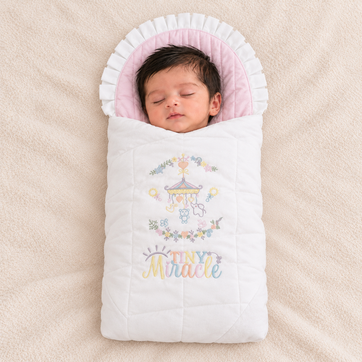 A sleeping newborn baby wrapped in a white quilted baby blanket with a pink ruffled hood, featuring a colorful embroidered carousel and the words 'TINY Miracle' on the front, laid on a soft beige blanket.