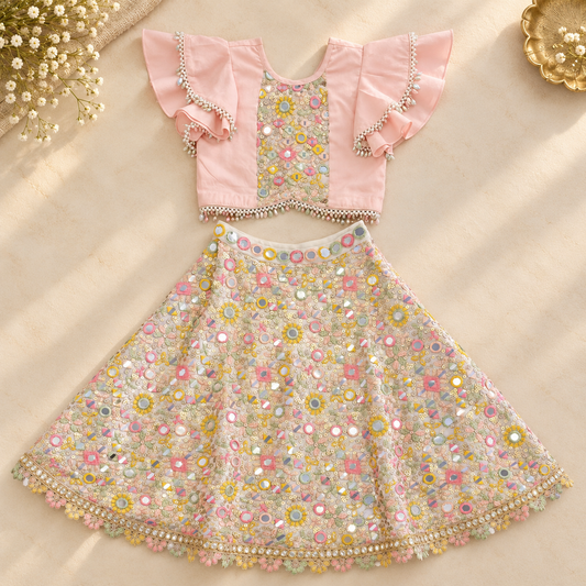 A two-piece girls' traditional outfit laid flat on a beige surface, featuring a light pink cropped top with ruffled sleeves and a heavily embellished floral skirt with mirror work and bead trim.