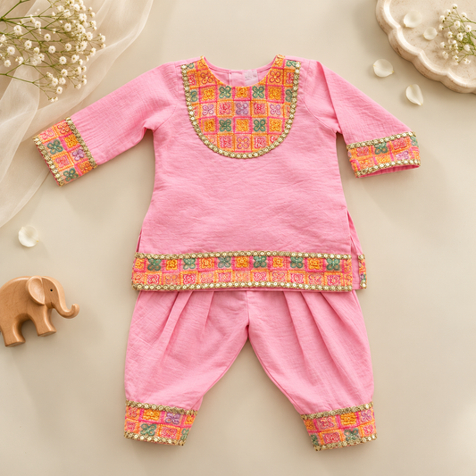 A baby boy's traditional Indian outfit consisting of a pink kurta and matching pants, decorated with colorful geometric embroidery and gold bead borders. The outfit is displayed in a styled flat-lay arrangement on a light beige background with decorative props including a wooden elephant, baby's breath, and a small dish.