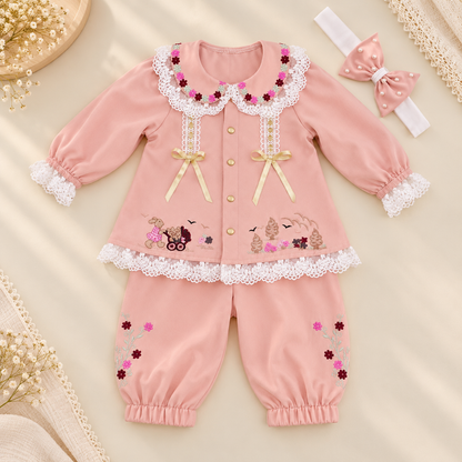 A baby girl's two-piece pink outfit set consisting of a long-sleeve button-up cardigan and matching pants, decorated with white lace, gold bows, and floral embroidery. The set is styled on a beige fabric background with a pink bow headband.