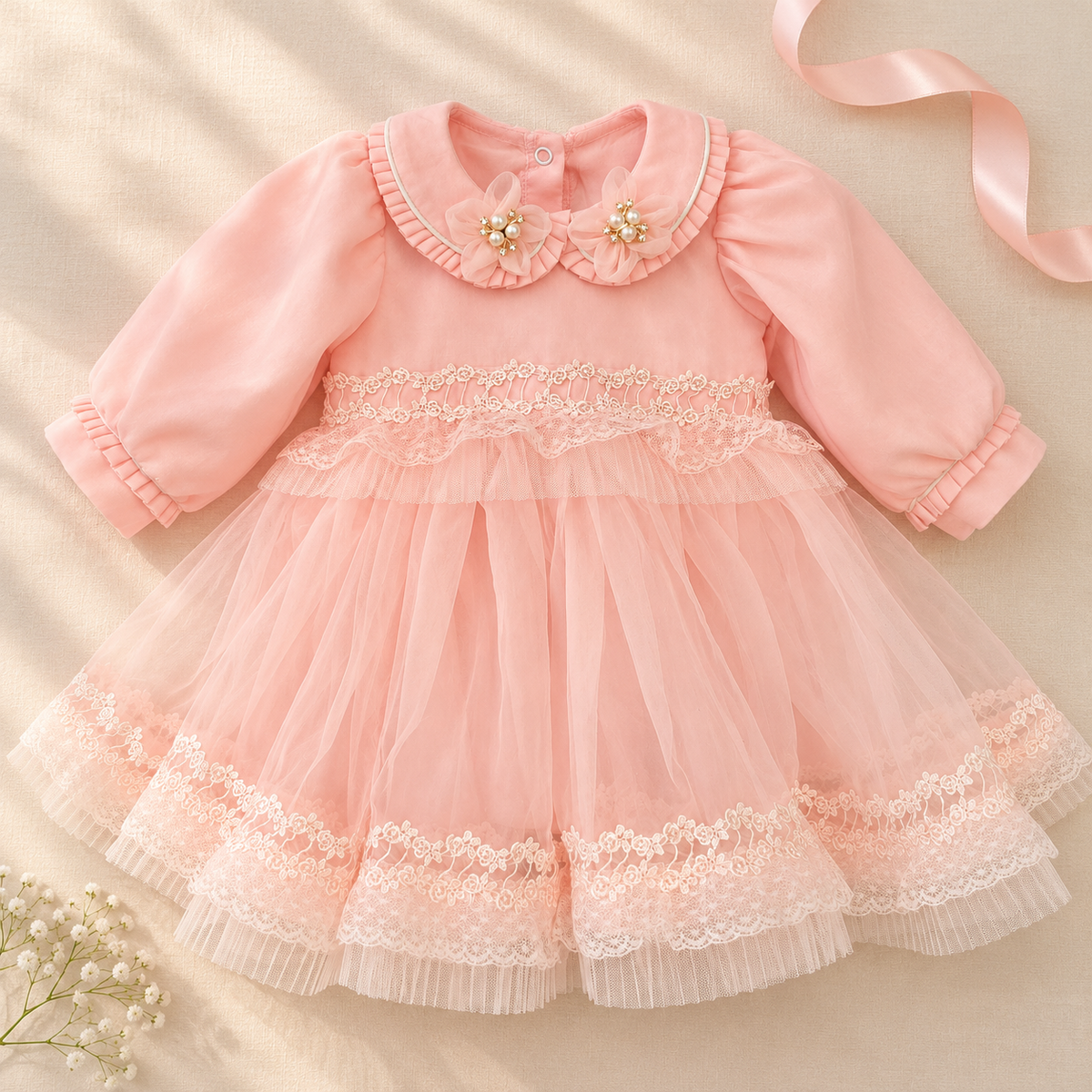 A baby girl's long-sleeve pink dress with a ruffled Peter Pan collar decorated with two pearl flower accents, a lace-trimmed bodice, and a layered tulle skirt with lace trim, laid flat on a beige fabric background with a pink ribbon and small white flowers.