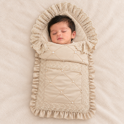A sleeping newborn baby wrapped in a beige quilted baby blanket with delicate lace trim and pearl-like buttons, lying on a soft beige fabric background.