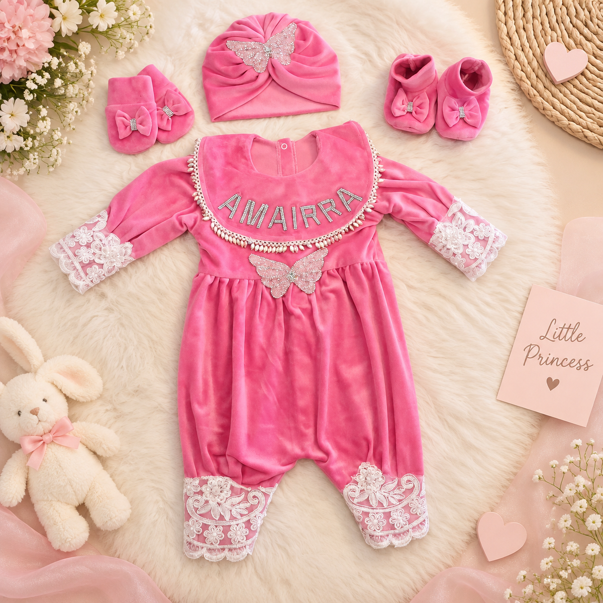 A pink baby girl's velvet one-piece jumpsuit with lace cuffs and hem, featuring the name 'AMAIRRA' in rhinestones and a butterfly applique. It is styled in a flat lay with a matching pink turban hat, booties, and accessories on a white faux fur background.