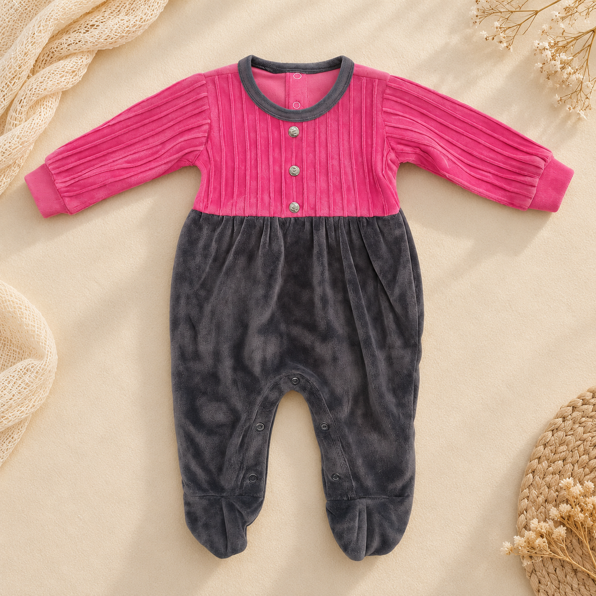 A baby footed one-piece romper in textured velvet, featuring a bright pink ribbed top and dark gray legs, laid flat on a beige fabric background with decorative props.