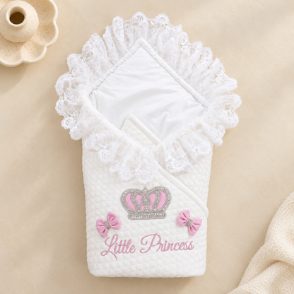 A white quilted baby wrap blanket with white lace trim, featuring a pink and silver crown design and the words 'Little Princess' in pink script. The blanket is displayed on a beige fabric background with a small ceramic candle holder prop.