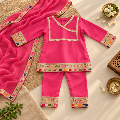 A bright pink traditional children's outfit set laid flat on a neutral surface, including a long kurta with embroidered multicolor borders, matching pants, and a draped dupatta. The outfit is styled with small decorative props such as flowers and a woven mat.