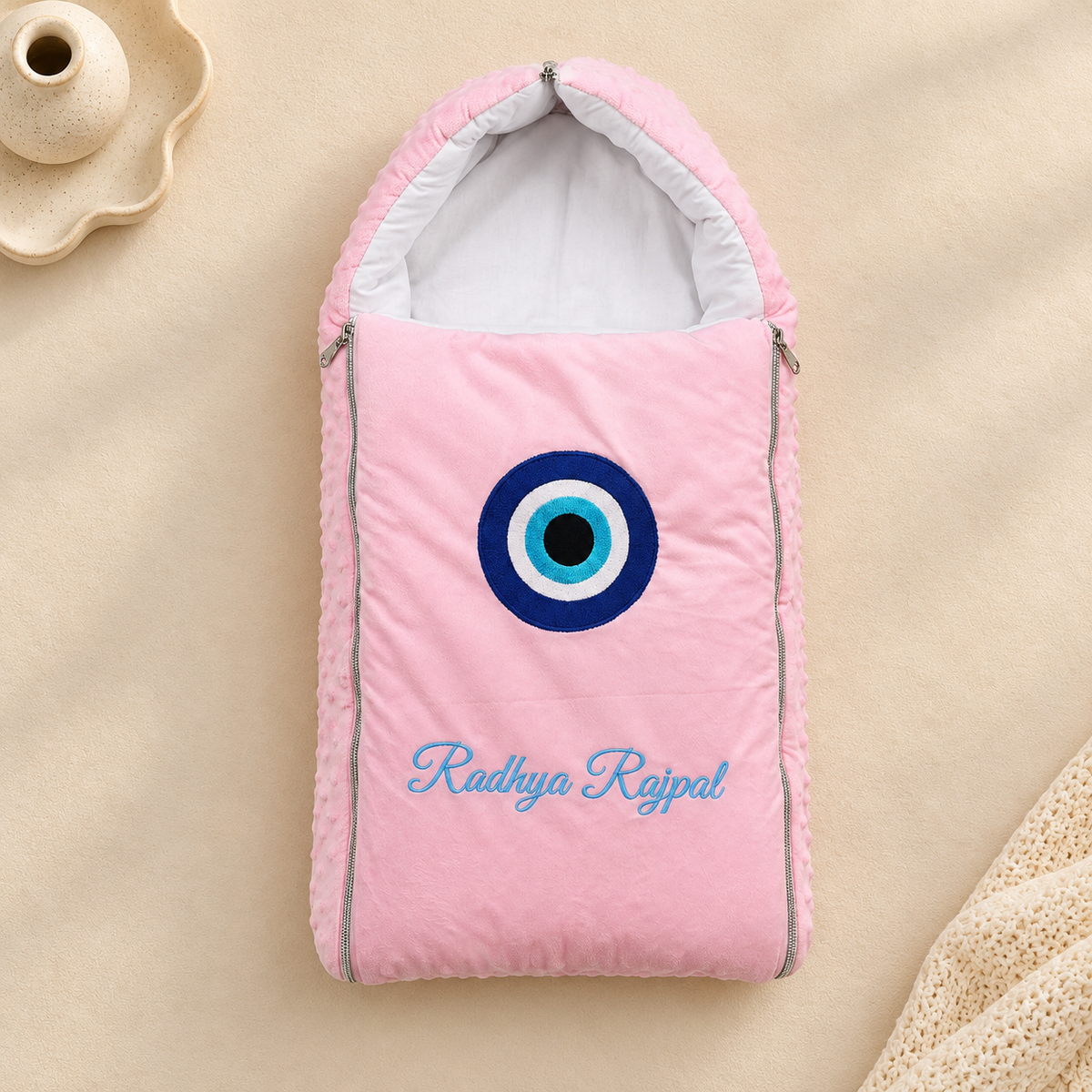 A pink baby footmuff or sleeping bag with a hood, featuring a blue and white evil eye emblem and the embroidered name 'Radhya Rajpal' in blue script. The product is laid flat on a beige fabric background with a small decorative ceramic object in the top-left corner.