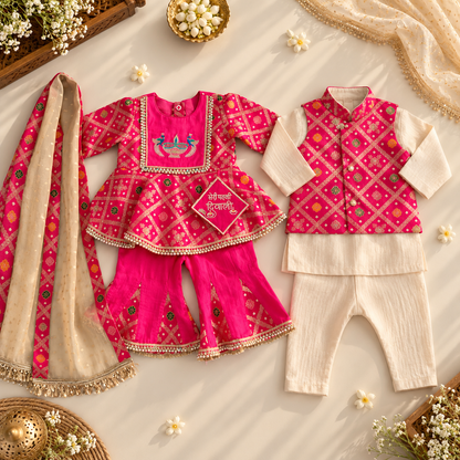 A styled flat-lay of a children's traditional outfit set on a cream fabric background with decorative flowers and accessories. The set includes a bright pink embroidered kurta with matching pants and a beige patterned dupatta, alongside a matching vest and cream pants.