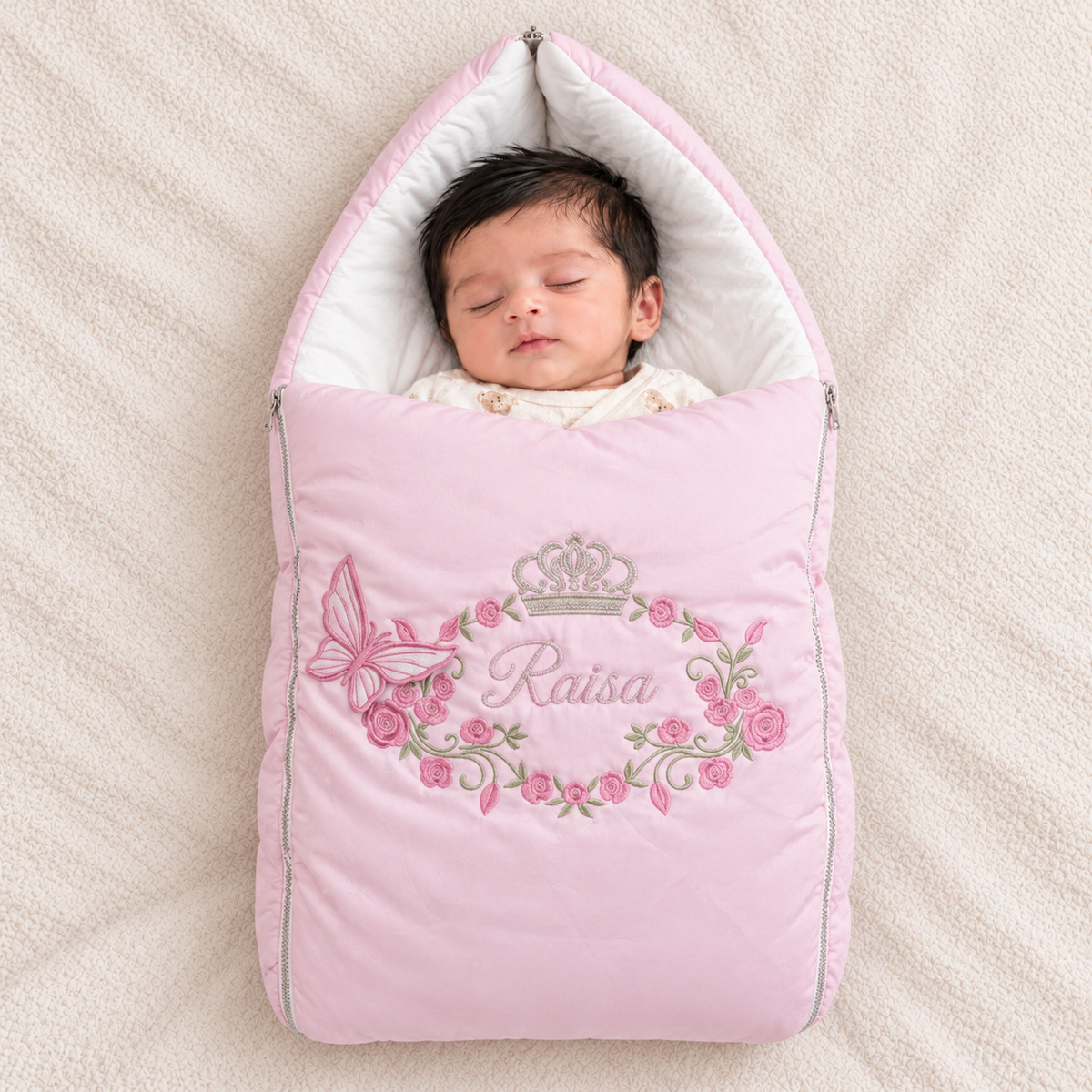 A sleeping newborn baby wrapped in a pink quilted footmuff bag with floral embroidery and the name 'Raisa' on a soft cream blanket.