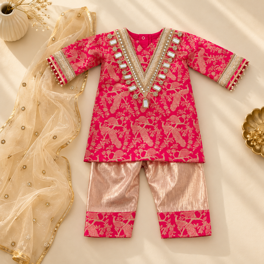 A baby boy's traditional Indian outfit laid flat on a cream fabric background, featuring a bright pink kurta with gold peacock motifs, a gold embellished V-neckline, and matching pink pants. A sheer beige dupatta with gold embroidery is draped to the left.