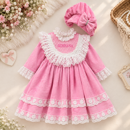 A pink baby dress with white lace trim, a matching pink bonnet with a bow, and the name 'GIRRANA' embroidered on the chest, arranged on a soft cream surface with decorative floral and wicker props.