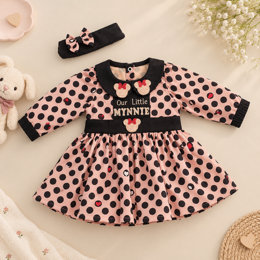 A baby girl's long-sleeve dress with a pink and black polka dot pattern, featuring a black velvet collar and cuffs, a matching black waistband, and Minnie Mouse-inspired embroidered details. A matching black and pink polka dot headband is placed above the dress on a beige fabric background with baby props including a plush bunny, flowers, and a woven mat.