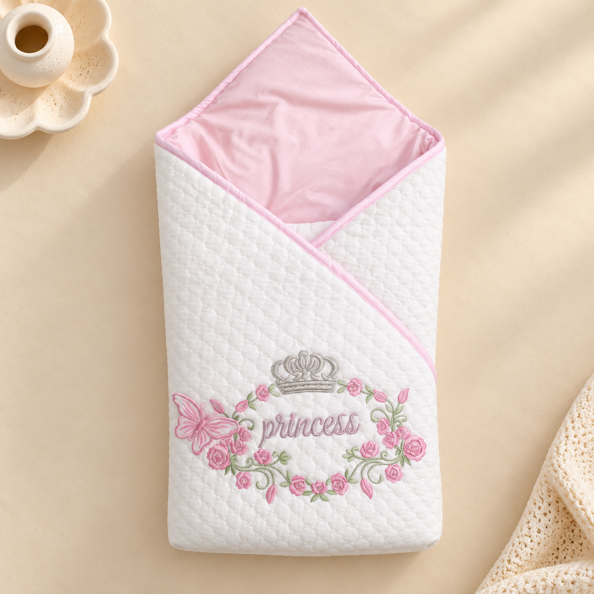 A white quilted baby swaddle blanket with a pink hood and pink trim, featuring an embroidered 'princess' design with a crown, flowers, and a butterfly. The blanket is displayed on a beige background with a small ceramic cup in the top left corner.