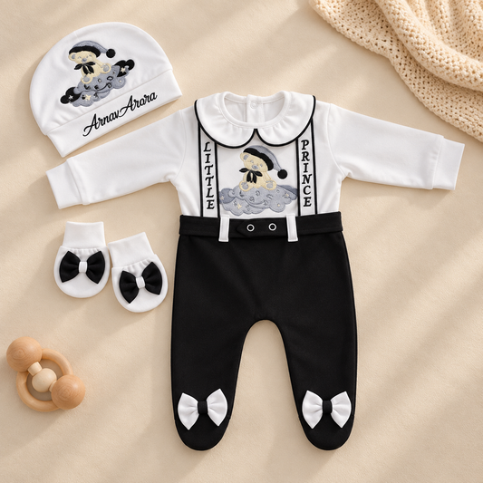 A baby outfit set laid flat on a beige fabric background, including a white long-sleeve footed romper with black suspenders and black pants, a matching beanie, and a pair of booties with black bow accents. A small wooden teething toy and cream knit fabric are also visible as styling props.