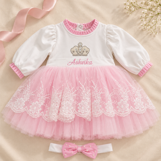 A baby girl's two-piece outfit set featuring a white long-sleeve top with pink ruffle trim, a pink tulle tutu skirt with white floral lace, and a matching pink polka-dot bow headband. The top is personalized with a silver crown graphic and the name 'Ashnika' in pink script.