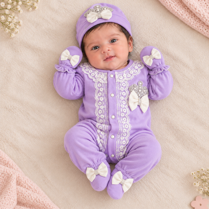 A baby lying on a soft beige blanket wearing a lavender purple footed onesie with white lace trim, white bows, and a matching lavender hat and mittens.