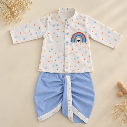 A baby clothing set laid flat on a warm beige background, featuring a white long-sleeve top with small blue and orange star prints and a rainbow patch on the chest, paired with light blue shorts with white trim.
