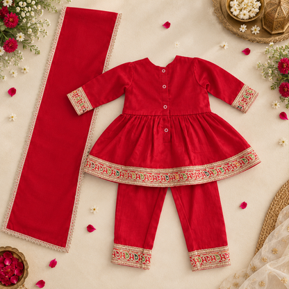 A baby girl's traditional Indian outfit set consisting of a red long-sleeve tunic with floral embroidered cuffs, matching red pants with matching cuffs, and a long red scarf with a beige lace border, arranged on a beige fabric background with decorative floral props.