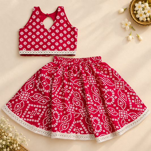 A two-piece baby girl's outfit set featuring a sleeveless red crop top with a white geometric bandhani pattern and a matching red flared skirt with a white bandhani print and lace hem, laid flat on a beige background with decorative flowers.