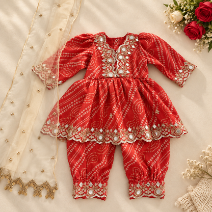 A baby girl's traditional Indian outfit consisting of a red printed kurta and matching pants with intricate silver mirror work and gold embroidery, styled with a cream dupatta and decorative flowers on a beige fabric background.