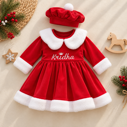 A baby girl's red velvet dress with a white faux-fur Peter Pan collar and cuffs, featuring the name 'Kridha' embroidered on the chest. The dress is styled with a matching red velvet beret hat, presented in a flat-lay arrangement with holiday-themed props on a beige fabric background.
