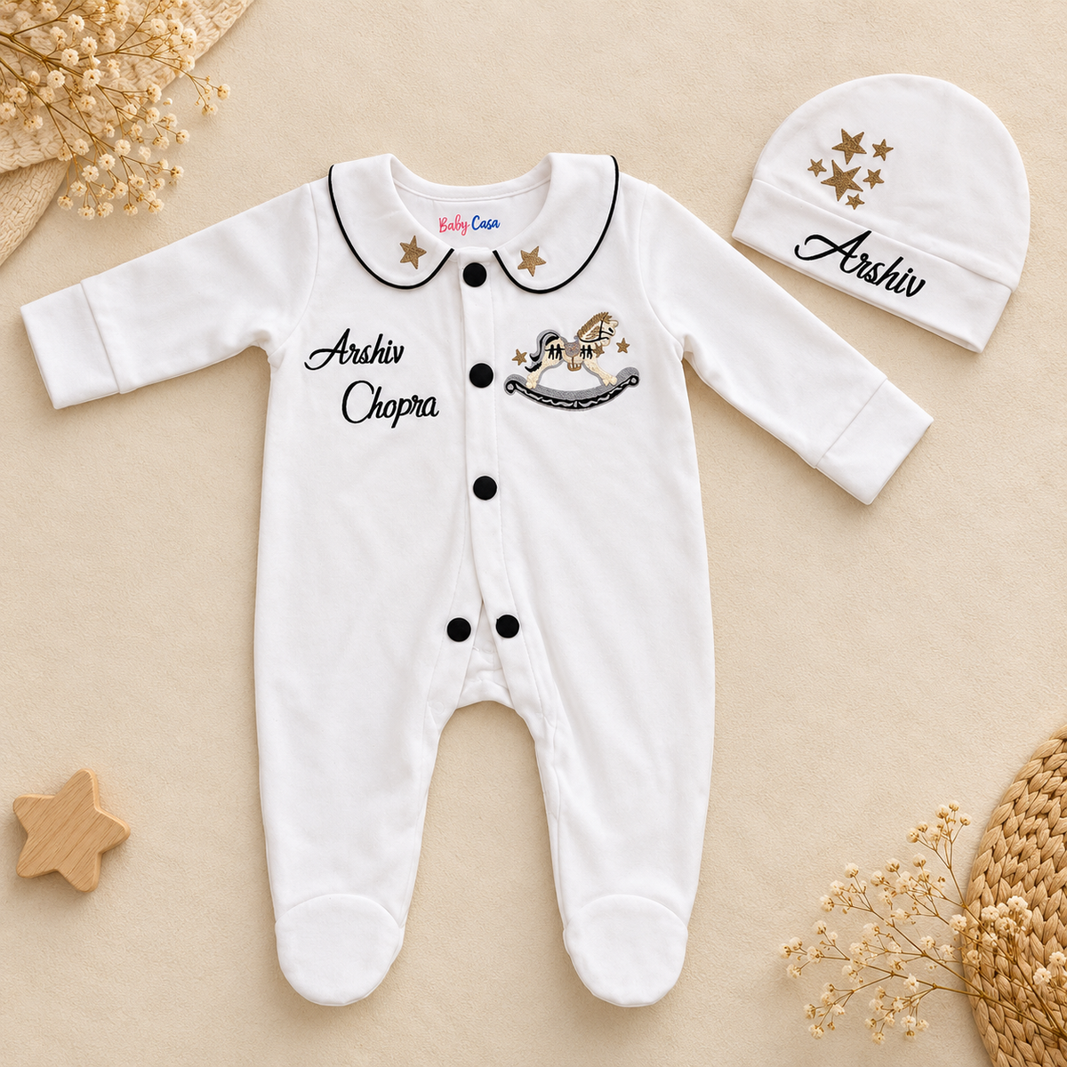 A white baby footed romper and matching beanie hat laid flat on a beige background. The romper has black trim around the collar, black buttons, black embroidered text reading 'Arshiv Chapra,' a small rocking horse graphic with gold stars, and 'Baby Crea' printed inside the collar.