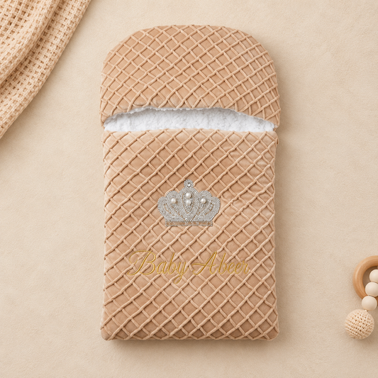 A beige textured baby pouch with a diamond-patterned exterior, a white fabric lining visible at the top, a silver crown graphic, and the words 'Baby Abar' in gold script, shown on a neutral beige background with styled props.