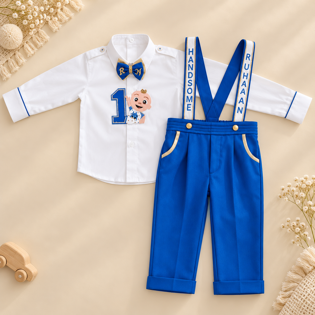 A baby boy's two-piece formal outfit set featuring a white long-sleeve shirt with a blue bow tie, number 1 patch, and 'Boy' text, paired with royal blue suspenders that read 'HANDSOME' and 'RUHAN'. The outfit is styled in a flat lay on a beige background with decorative props.