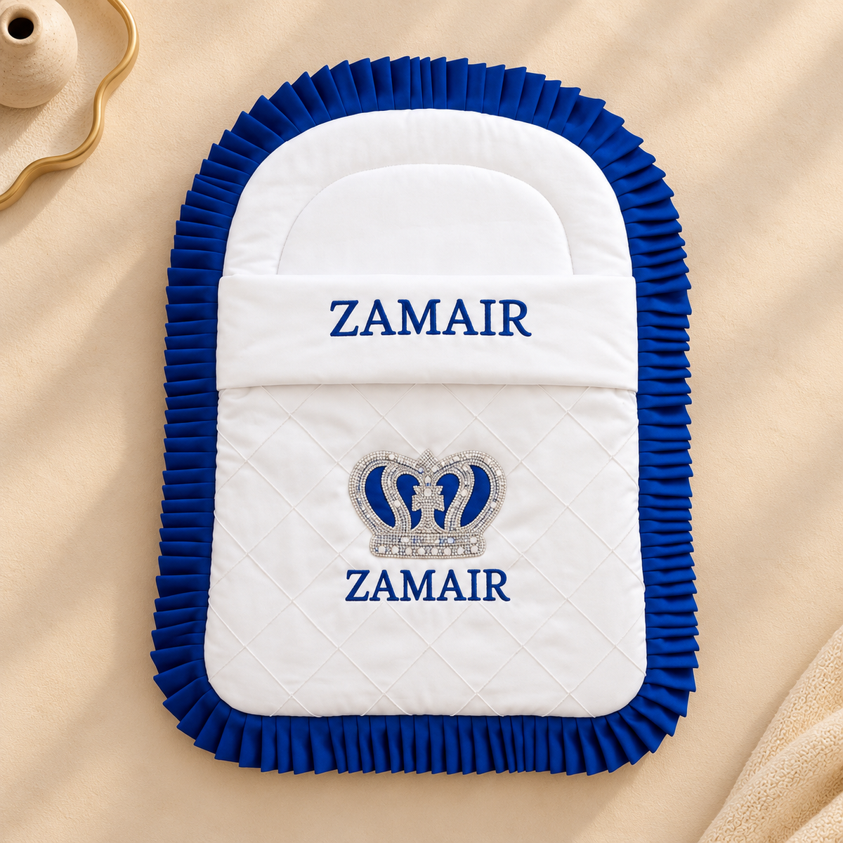 A white quilted baby blanket or footmuff with a royal blue ruffled border, featuring the brand name 'ZAMAIR' embroidered in blue on the front, along with a silver and blue crown motif. The product is laid flat on a beige fabric background.