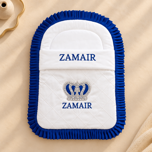 A white quilted baby blanket or footmuff with a royal blue ruffled border, featuring the brand name 'ZAMAIR' embroidered in blue on the front, along with a silver and blue crown motif. The product is laid flat on a beige fabric background.