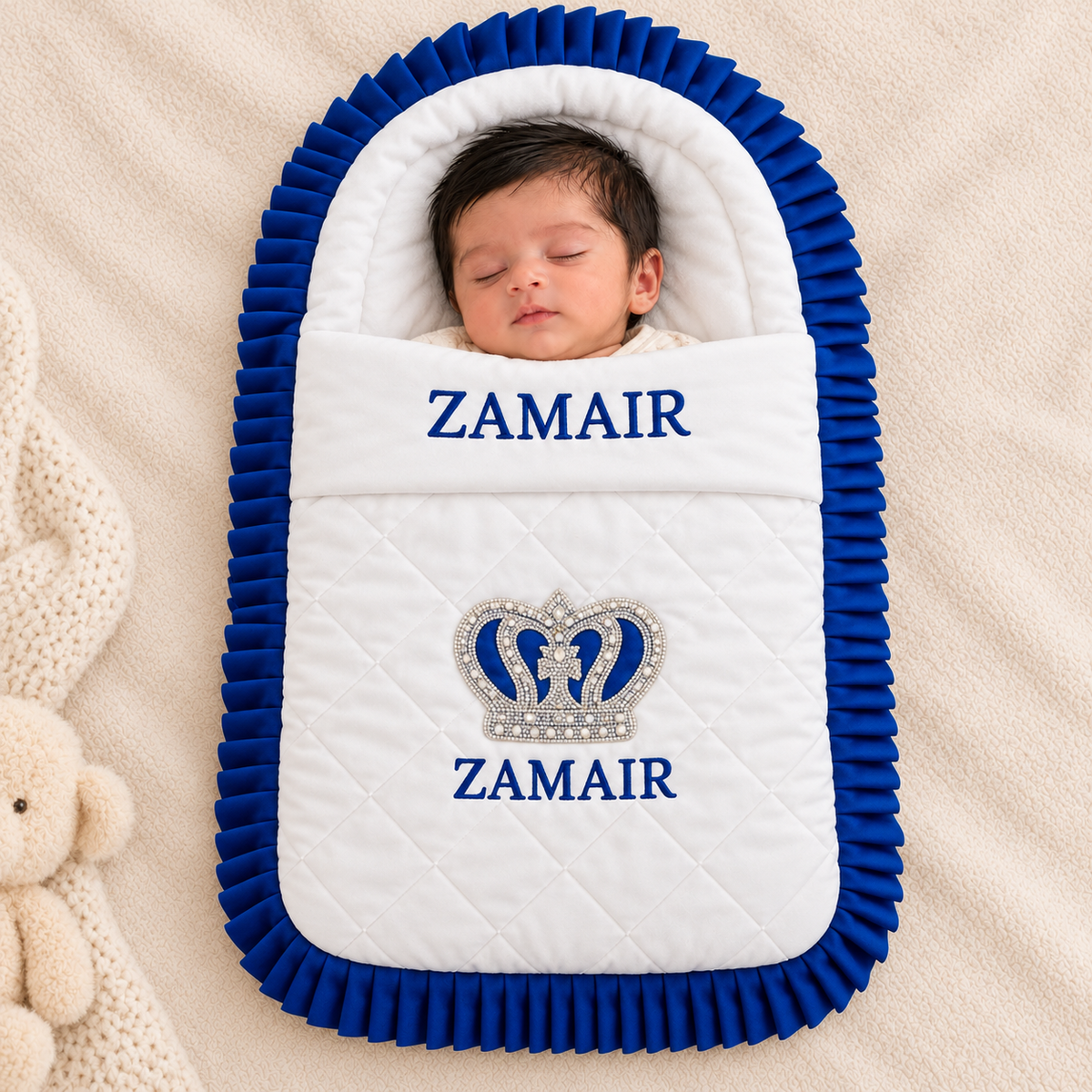 A sleeping newborn baby wrapped in a custom quilted white and royal blue swaddle blanket with pleated trim. The blanket features the name 'ZAMAIR' embroidered in blue on both sides and a rhinestone-embellished crown motif in the center.