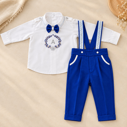 A baby boy's formal outfit set consisting of a white long-sleeve shirt with a blue bow tie and a floral monogram 'A' on the chest, paired with royal blue pants featuring light blue patterned suspenders, laid flat on a beige fabric background.