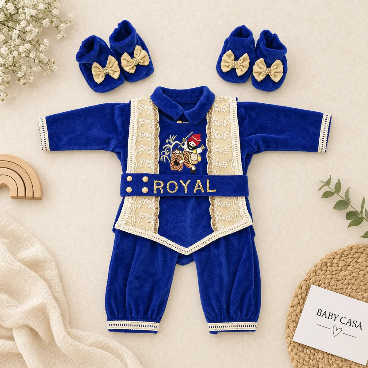 A baby royal-themed velvet outfit set in royal blue, featuring a long-sleeve top and pants with cream lace trim, a matching belt with gold buttons and 'ROYAL' text, and matching booties with bow accents, arranged on a neutral fabric background with decorative props.