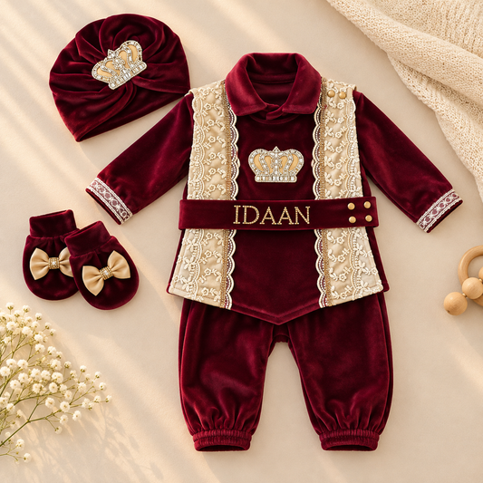A baby boy's three-piece royal velvet outfit set in deep burgundy, featuring a long-sleeve top with lace-trimmed collar and sleeves, matching pants, a decorative waist belt with gold lettering, a crown applique, a headband with a crown, and matching bowie booties, arranged on a beige fabric background with baby accessories.