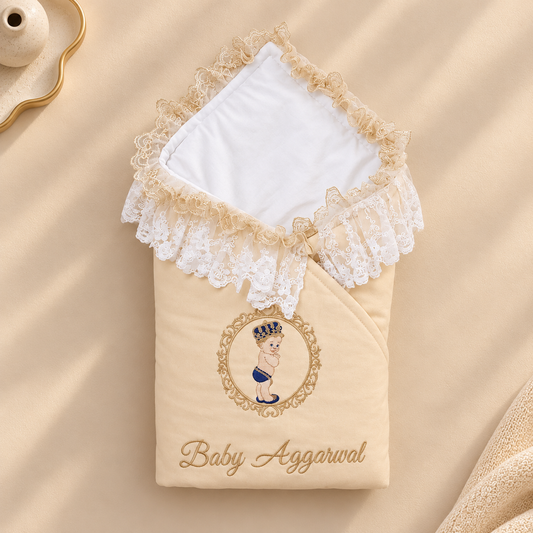 A beige baby swaddle wrap with a white hooded lining and beige lace trim, featuring an embroidered portrait of a baby in a crown and the text 'Baby Aggarwal' on the front, styled on a beige fabric background.