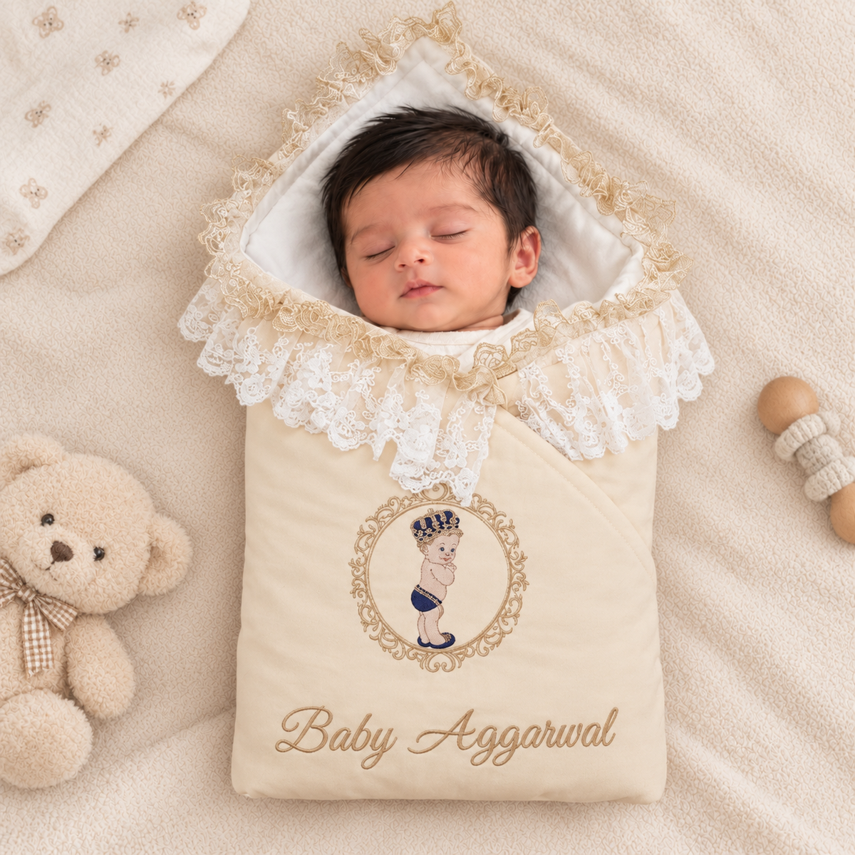 A sleeping newborn baby wrapped in a cream-colored personalized baby swaddle blanket with white lace trim, featuring an embroidered illustration of a baby in a crown and the text 'Baby Aggarwal'. The swaddle is styled in a flat-lay arrangement with a teddy bear and a wooden rattle on a soft beige blanket background.